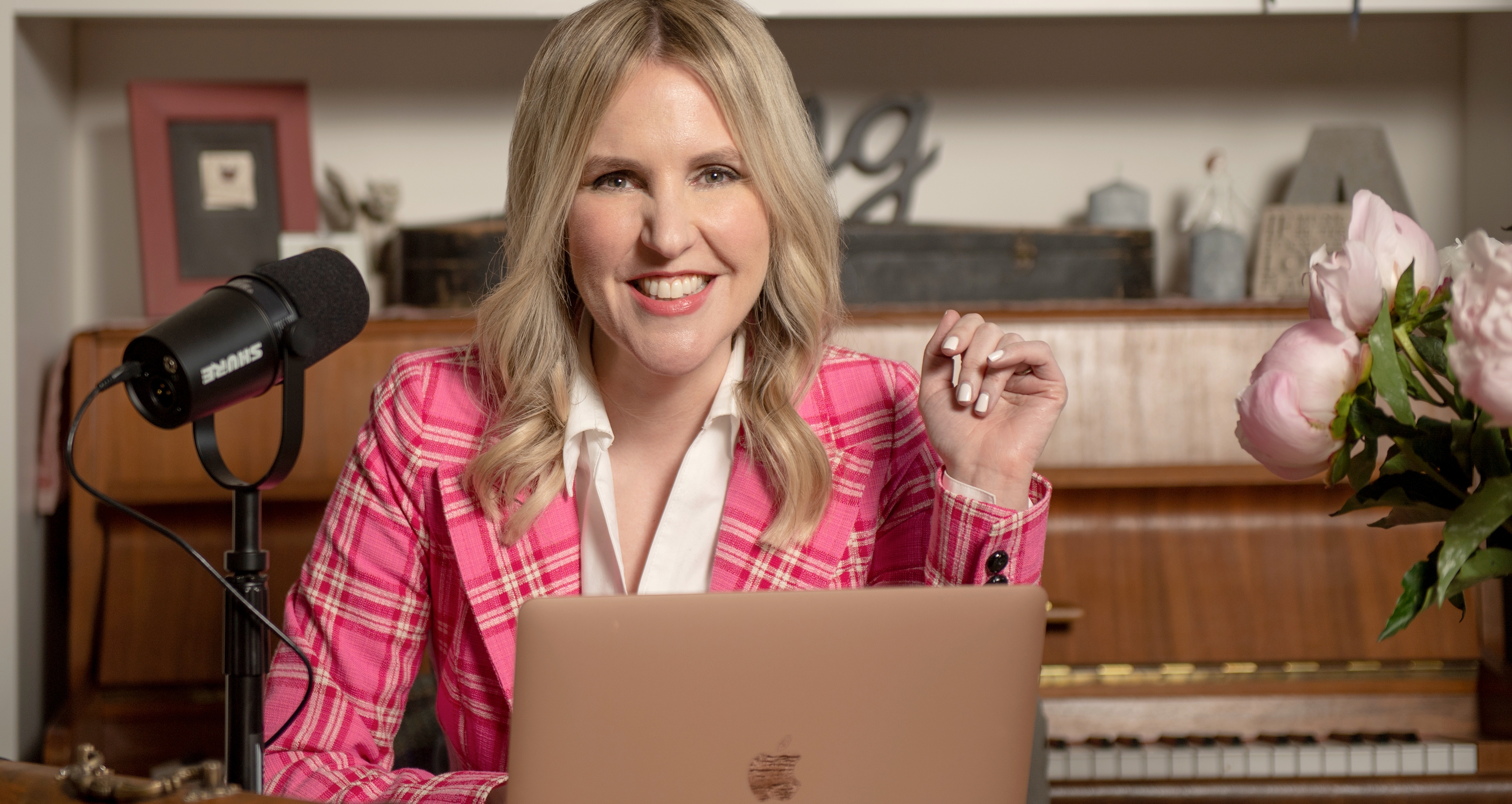 Ardra Shephard smiles into the camera. She is sitting behind an open laptop, a microphone to her right.