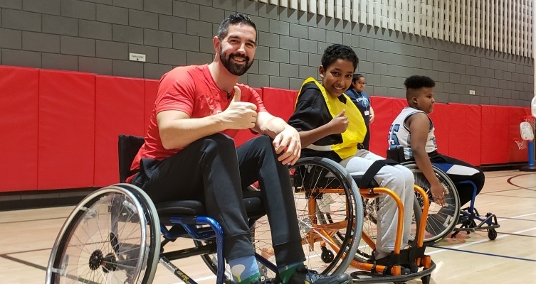 Two people, using wheelchairs, smile into the camera, with thumbs up.