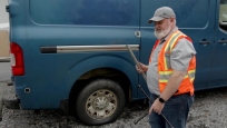 A man stands next to a van. The van has a flat tire.