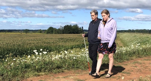 A man and woman walk along a dirt path. The man is using a white cane.