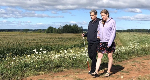 A man and woman walk along a dirt path. The man is using a white cane.